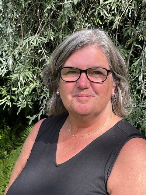 Sophie Sowerby, an Organisation Development practitioner and coach, stands outdoors in front of leafy green foliage. With grey hair and glasses, she wears a black sleeveless top and smiles slightly at the camera.